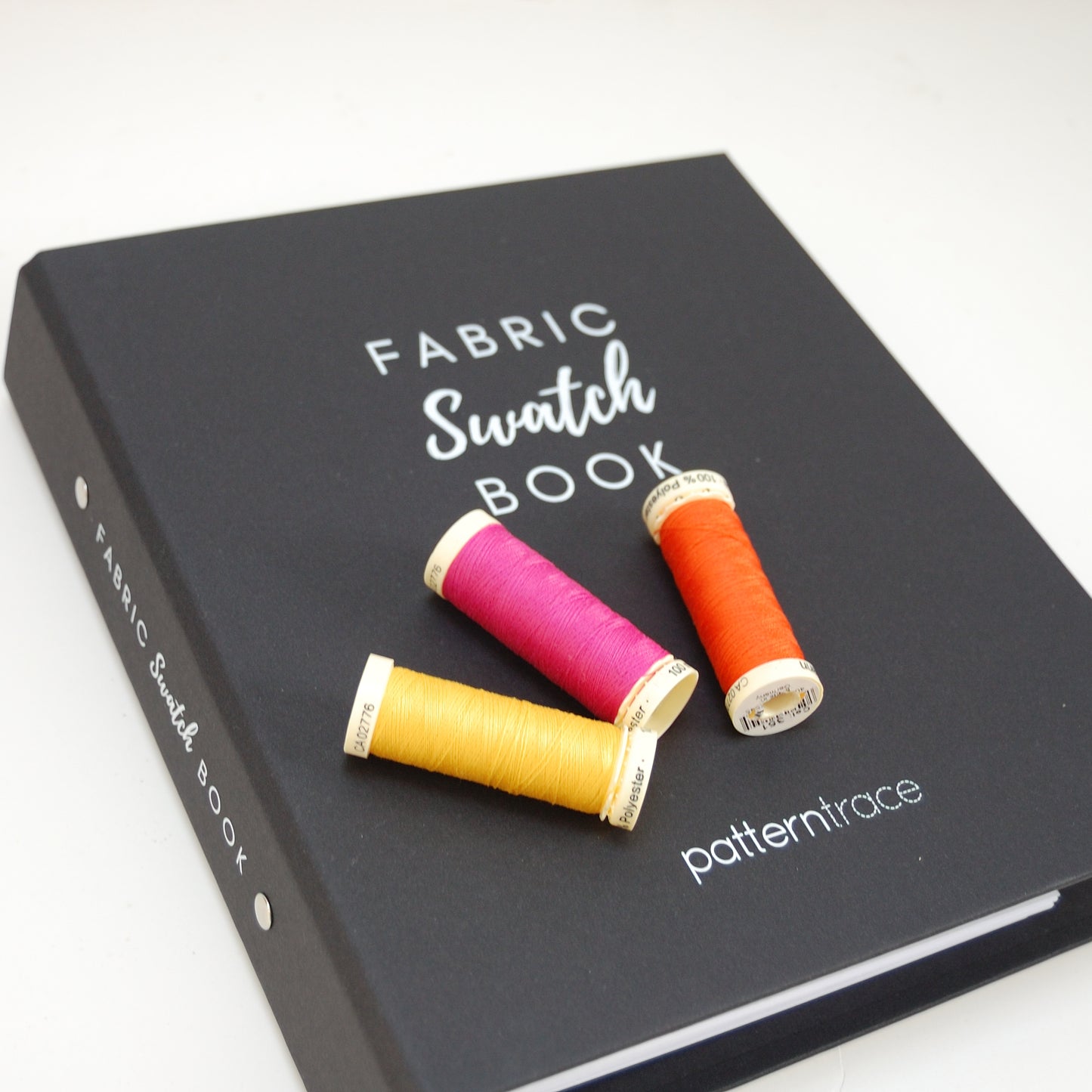 A black binder that says Fabric Swatch Book and Patterntrace in white letters. There are three spools of thread on the book. From the left the spools are yellow, fuscia and orange