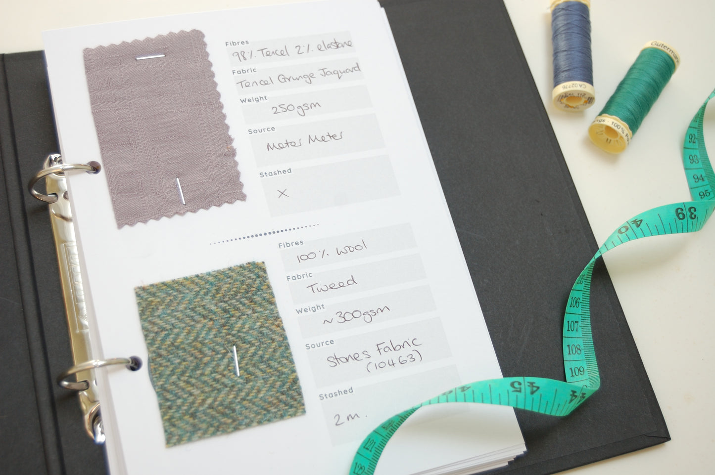 an open binder with two swatches of fabric one is taupe and one is green. There is a teal tape measure and two spools of thread to the right one is blue and the other is green