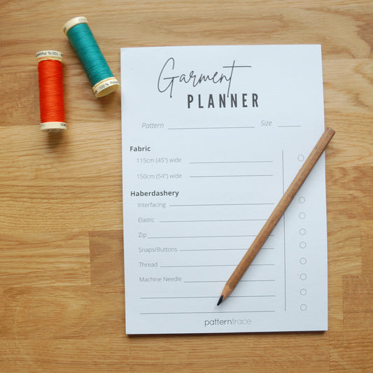 A note pad sitting on an oak table top.  It says Garment Planner and has lines for planning haberdashery needed for a garment.  There is a wooden pencil on the lower right side and two spools of thread on the left, one orange and one teal