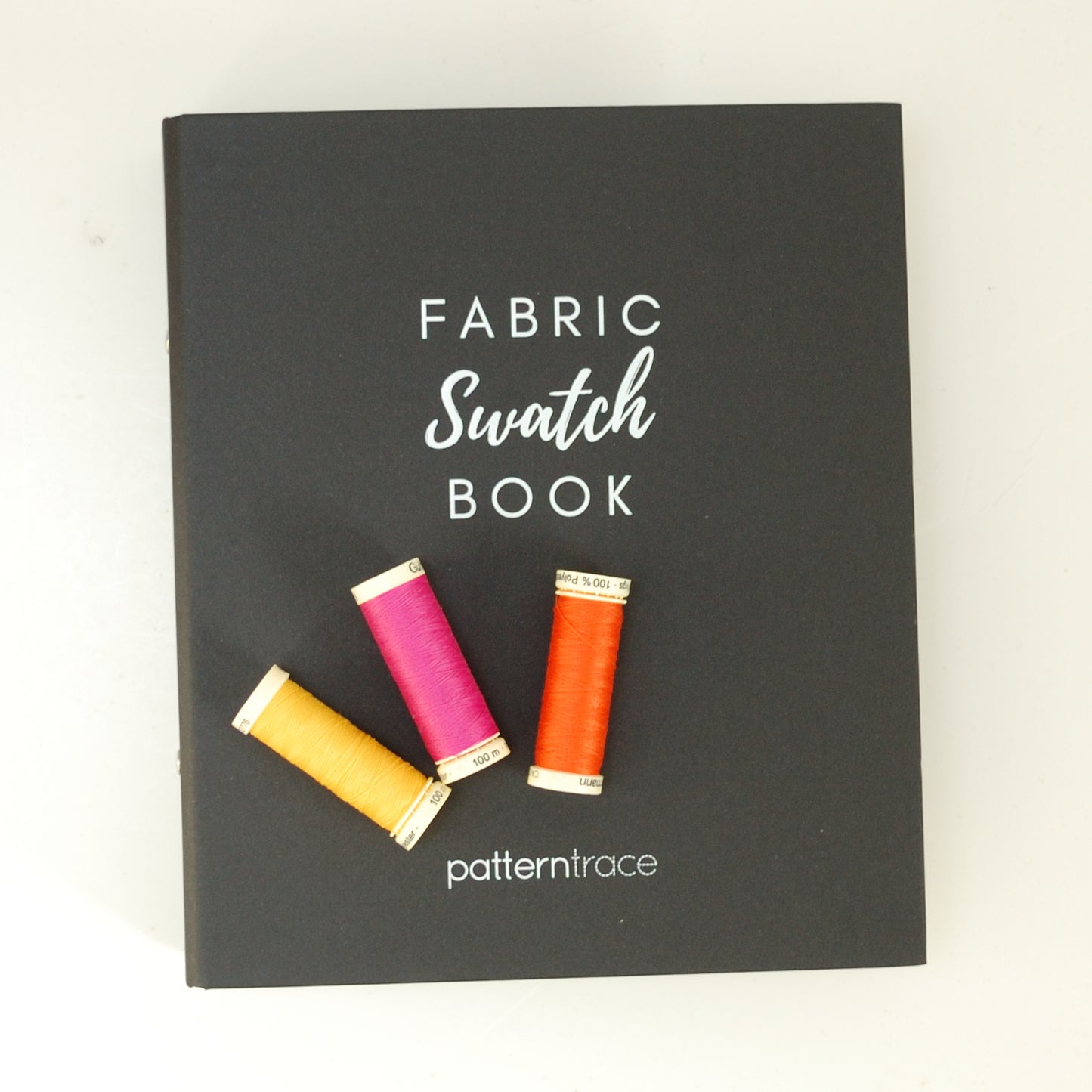 A black binder that says Fabric Swatch Book and Patterntrace in white letters. There are three spools of thread on the book. From the left the spools are yellow, fuscia and orange