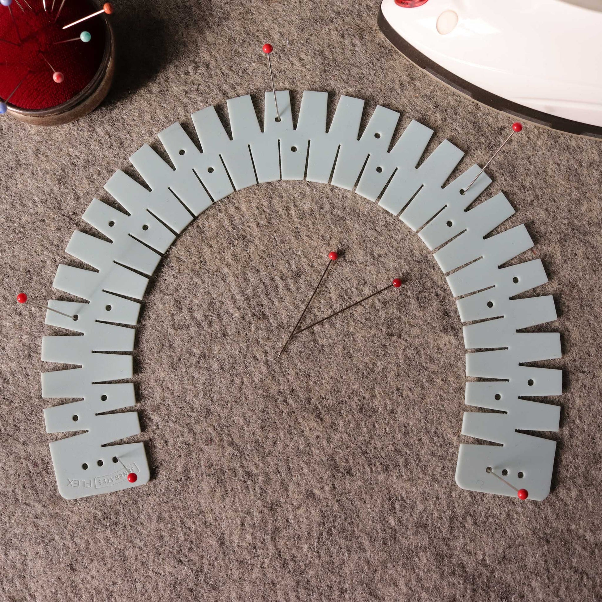 A gray flexible silicone hemmer in the shape of an upside down U on a gray wool mat with sewing pins with red tops