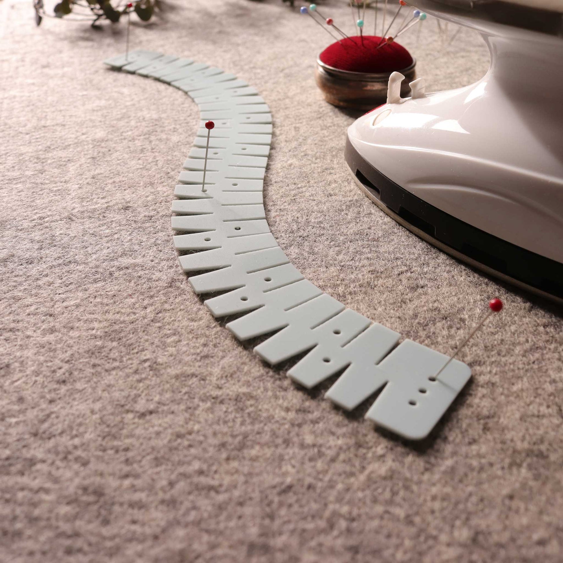 A gray flexible silicone hemmer in a reverse s curve on a gray will matt with a mini iron and a red pin holder with some pins