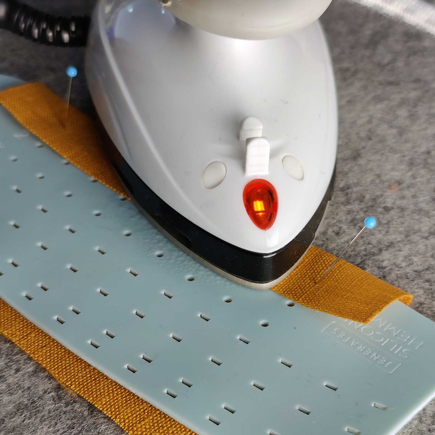 A teal colored silicone hemmer with some orange fabric being ironed with a mini iron