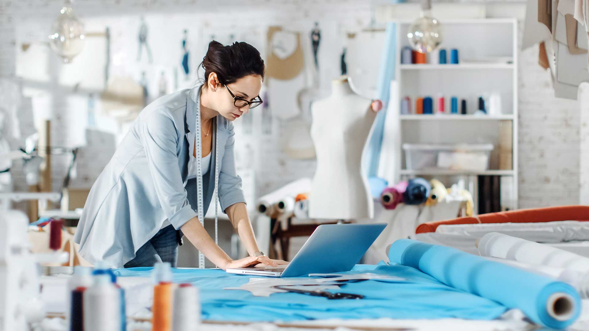 A white themed sewing studio with a lady looking at a laptop. There are bolts of fabric in blue and white.