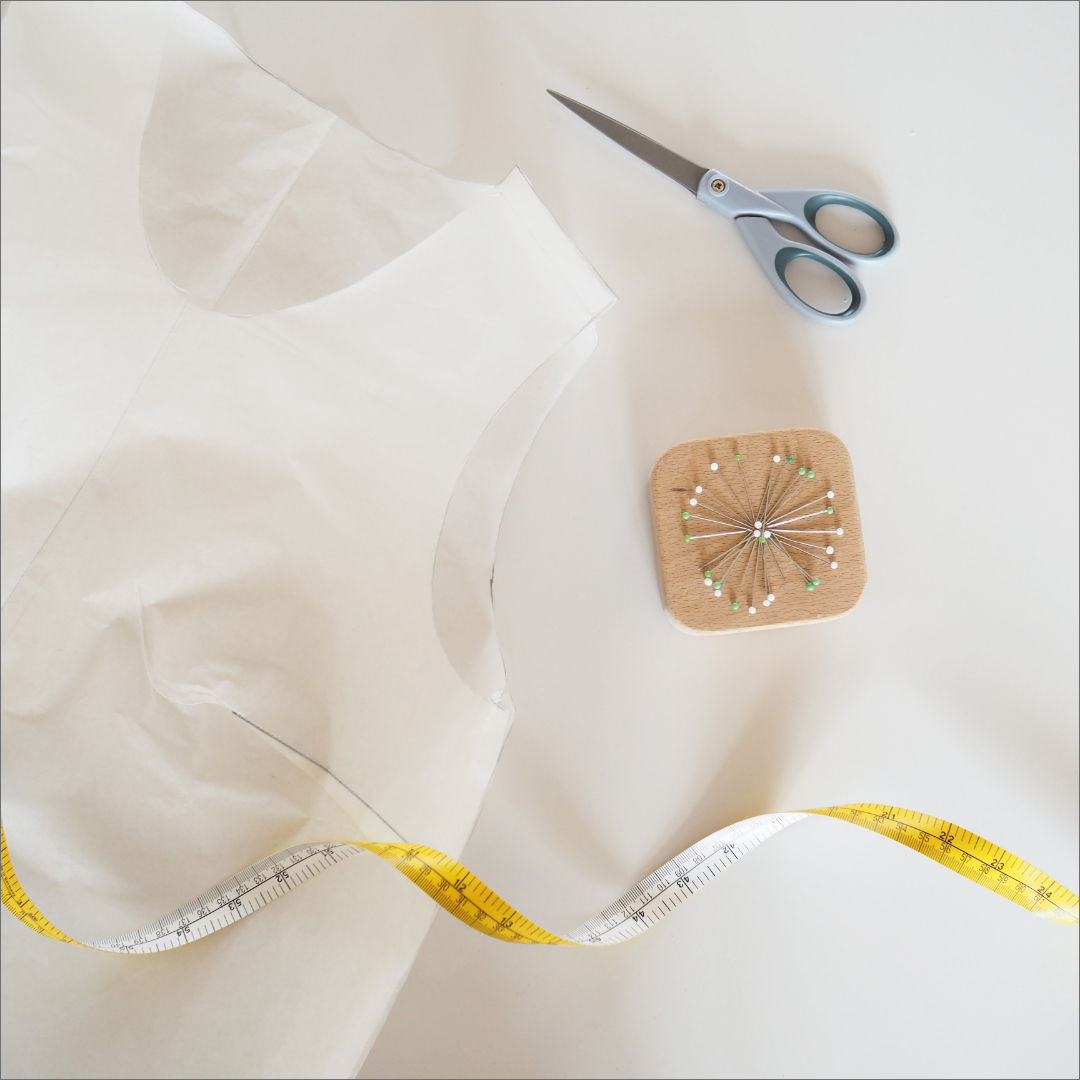 a bodice sloper sewn from patterntrace paper.  There is a white and yellow tape measure along the bottom, a pair of blue handled scissors on the top right and a corkboard with pins below the scissors