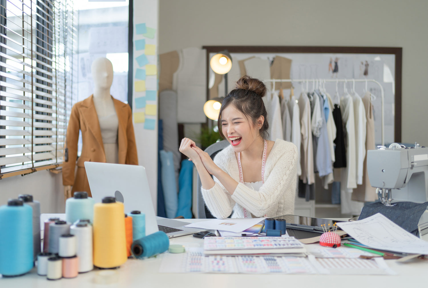 A pretty Asian teenager sitting in a sewing room smiling as she is looking at her open laptop