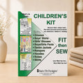 A sewing kit box for children's clothing by Sure Fit Designs, standing upright on a wooden surface with a soft-focus warm indoor background. The box is white and green with large text reading 'CHILDREN'S KIT' and 'FIT then SEW', plus a list of garment types and a small product photo panel on the left.