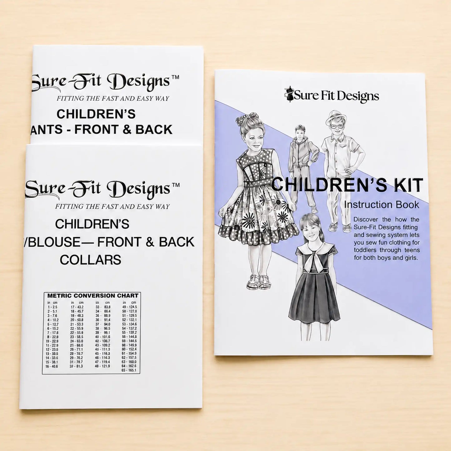 Two printed sewing pattern booklets from Sure Fit Designs laid on a light wood surface. One booklet shows children's pants front and back, and the other shows a children's kit instruction book cover with sewing illustrations and text.