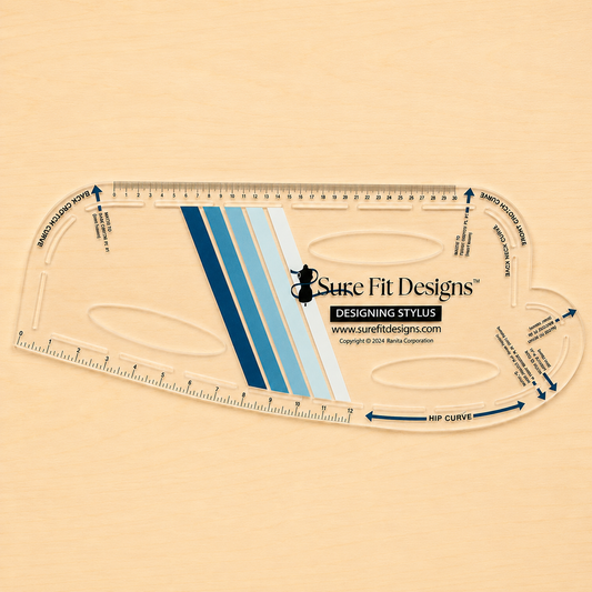 A clear high grade plastic measuring tool labeled Sure Fit Designs Designing Stylus, shown on a light beige wood background. The tool includes printed measurement markings, blue and white diagonal stripes, and printed guide text and arrows.