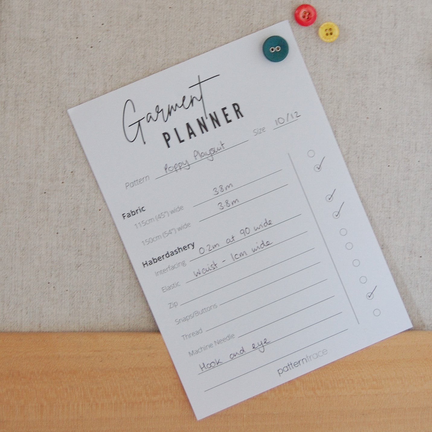 A note pad sitting on an oak table top.  It says Garment Planner and has lines for planning haberdashery needed for a garment.  There are three buttons on the upper right corner, teal, orange and yellow