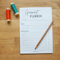 A note pad sitting on an oak table top.  It says Garment Planner and has lines for planning haberdashery needed for a garment.  There is a wooden pencil on the lower right side and two spools of thread on the left, one orange and one teal