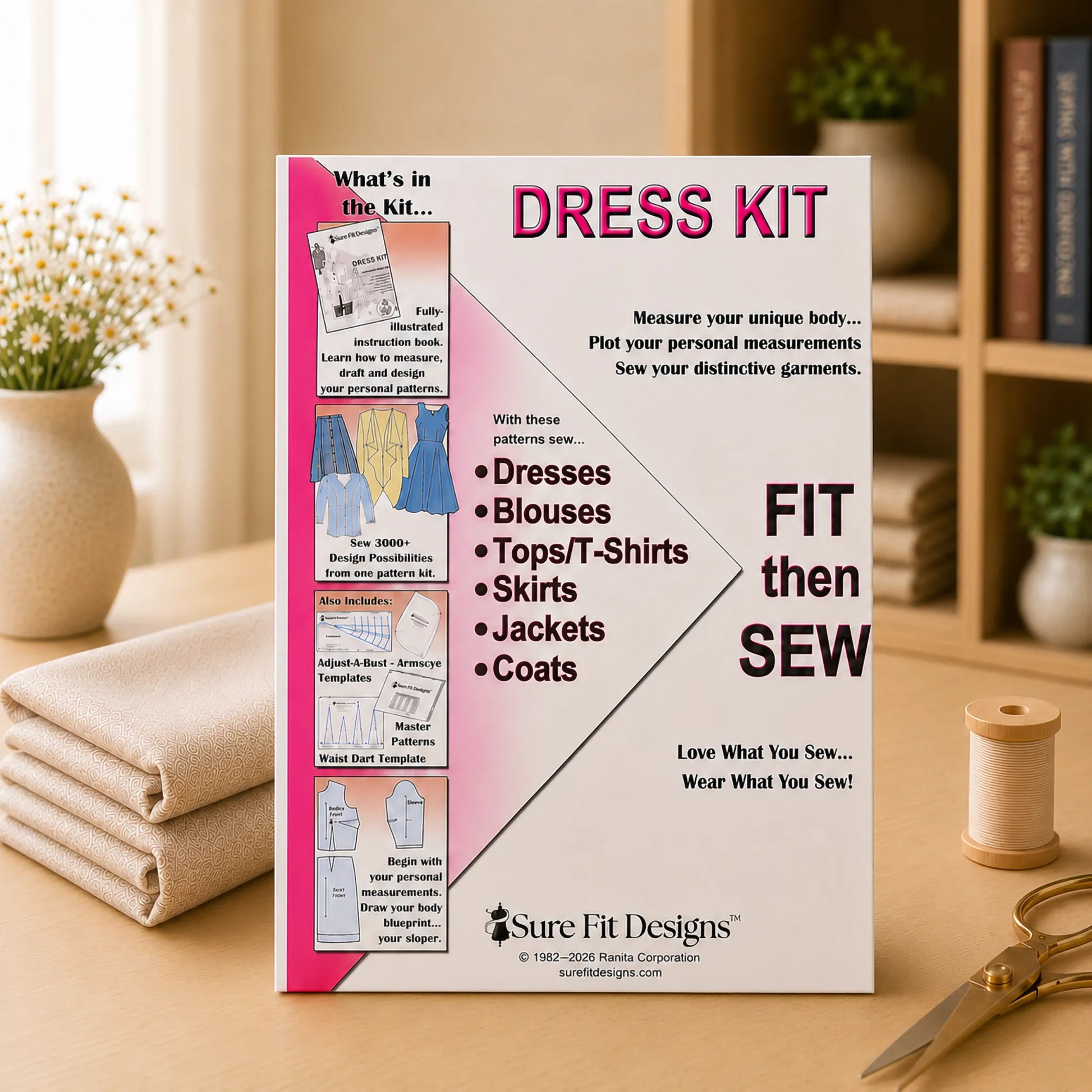 A sewing instruction booklet titled 'Dress Kit' by Sure Fit Designs, standing upright on a wooden table with a spool of thread and scissors nearby. The booklet features a pink and white cover with text and illustrations showing different garment types.
