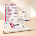 A sewing dress kit set displayed on a light wood surface, including a printed instruction booklet, a drafting course card, a curved measuring ruler, and a roll of white fabric.