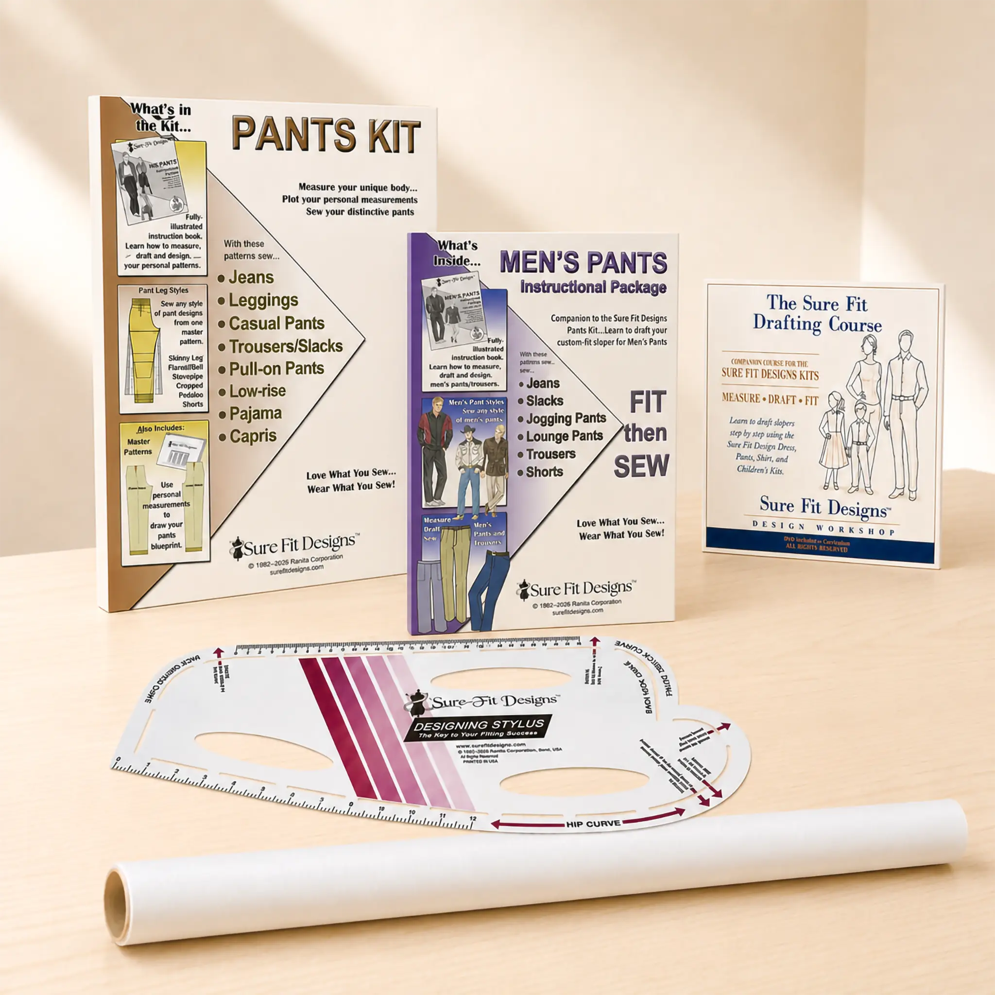 Sure Fit Designs pants fitting kit displayed on a light wood surface with a beige background. The kit includes instruction booklets, a pattern transfer sheet, a curved measuring template, and a rolled transfer sheet.