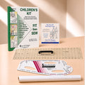A children's sewing kit laid out on a light wood surface, including a green and white instruction booklet, a small blue-and-white guide card, a clear gridded quilting ruler, a black rotary cutter, a white curved measuring template, and a rolled white paper sheet. Visible branding includes Sure Fit Designs and The Save Fit Designs.