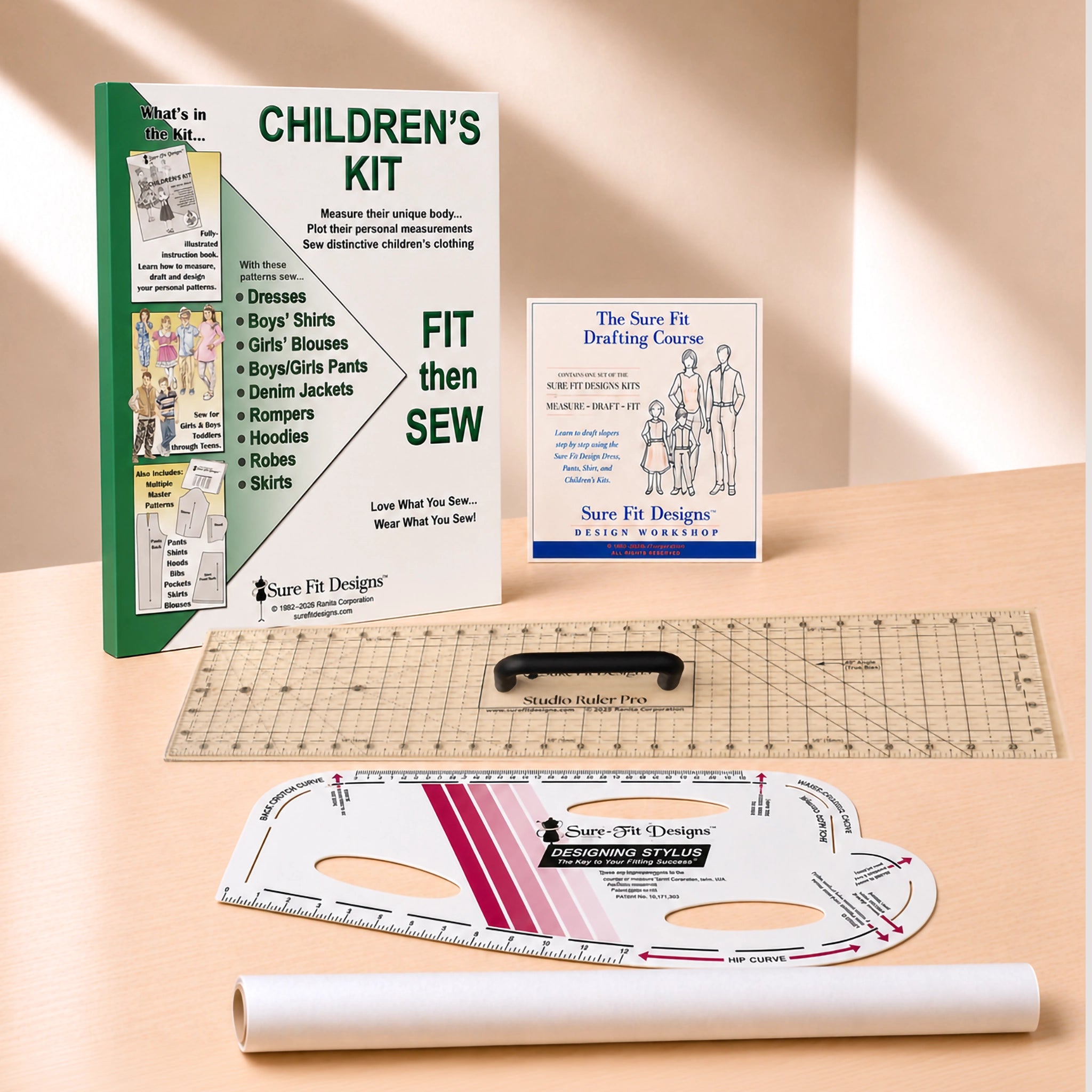 A children's sewing kit laid out on a light wood surface, including a green and white instruction booklet, a small blue-and-white guide card, a clear gridded quilting ruler, a black rotary cutter, a white curved measuring template, and a rolled white paper sheet. Visible branding includes Sure Fit Designs and The Save Fit Designs.