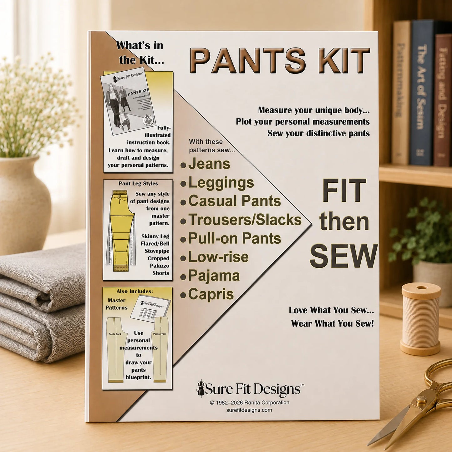 A 'Pants Kit' by Sure Fit Designs by Lori Fitzgerald is displayed on a wooden table in a styled home environment with a sewing book, thread spool, and scissors. The kit packaging features a white and tan design with illustrations of different pant styles.