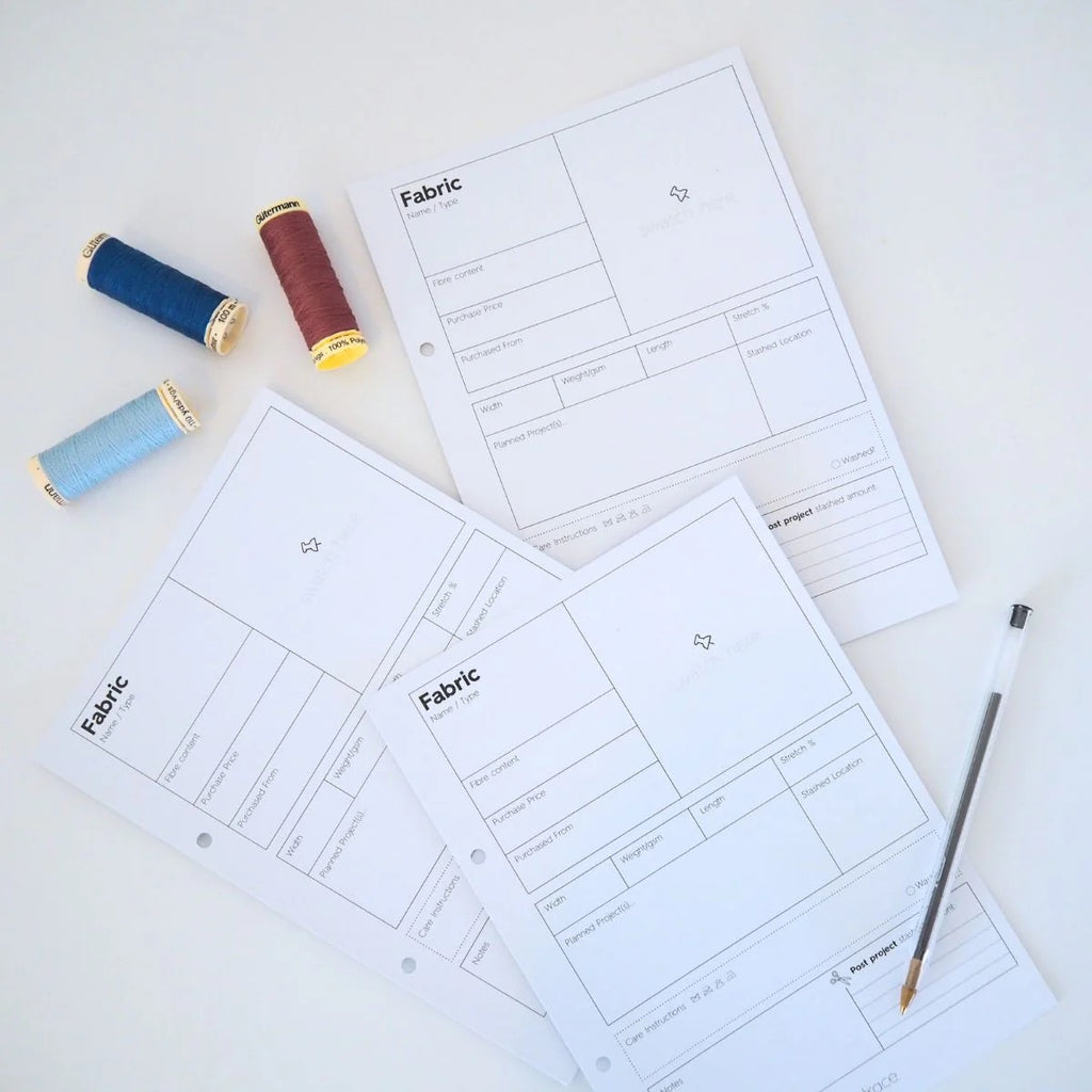 blank pages that are templates for pages for describing fabric qualities.  There are blue and red spools of thread in the upper left corner and a black pen in the lower right corner