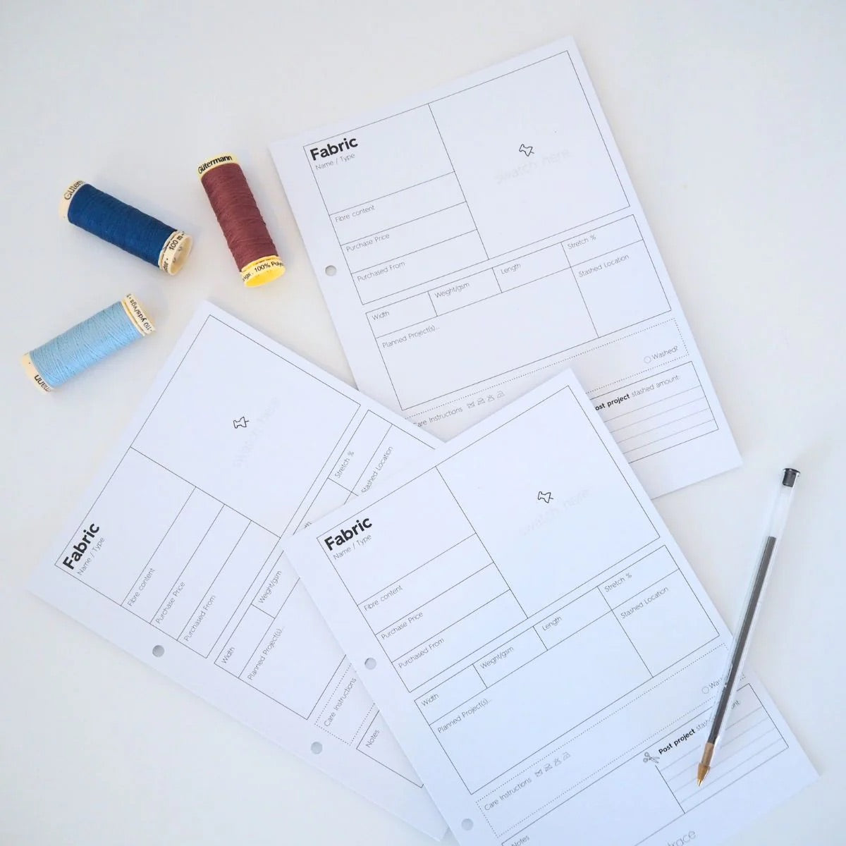 blank pages that are templates for pages for describing fabric qualities.  There are blue and red spools of thread in the upper left corner and a black pen in the lower right corner
