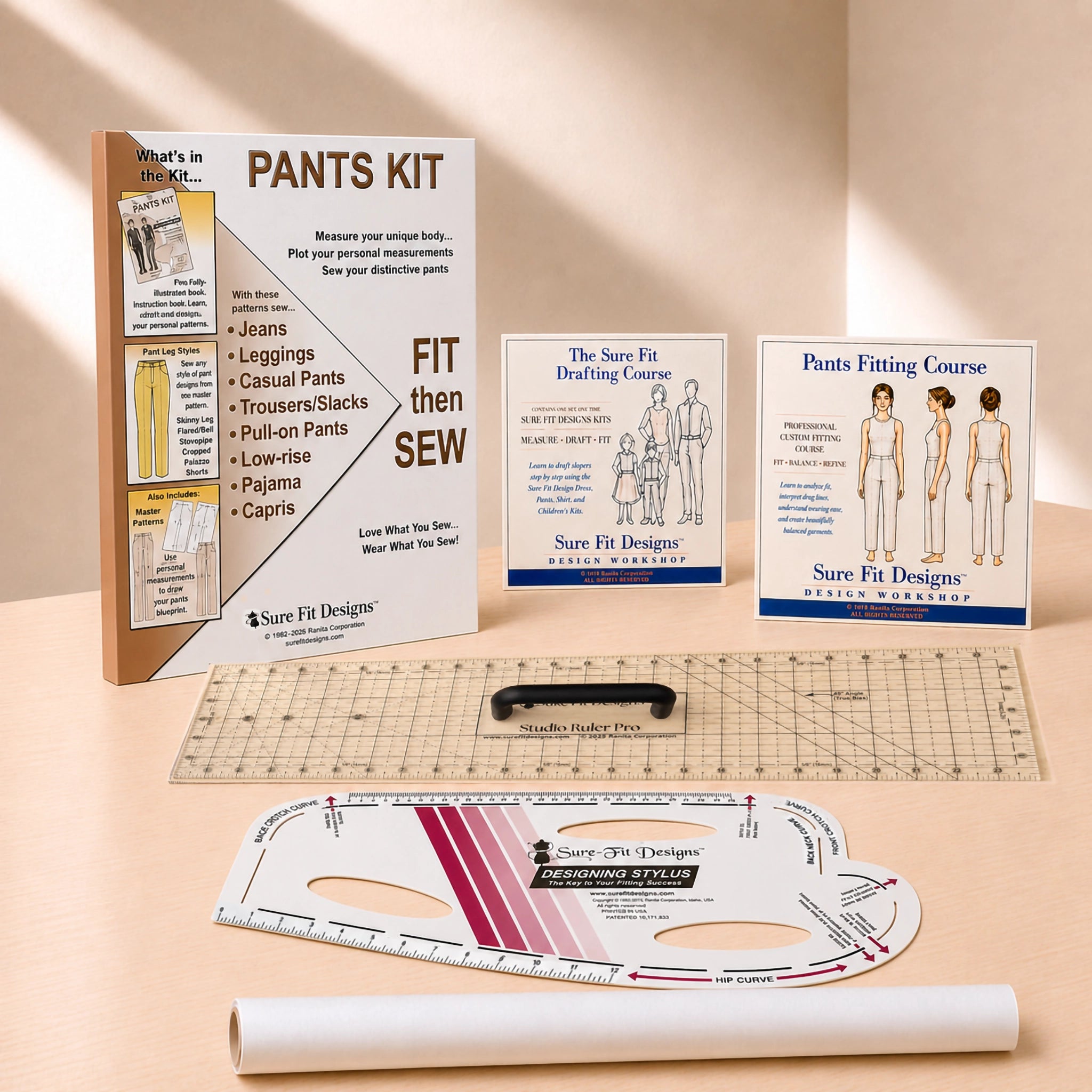 A sewing pattern kit and fitting tools arranged on a warm beige studio surface. Visible items include a 'PANTS KIT' instruction booklet, two Sure Fit Designs course cards, a clear acrylic ruler, a curved measuring template, and a white fabric roll.