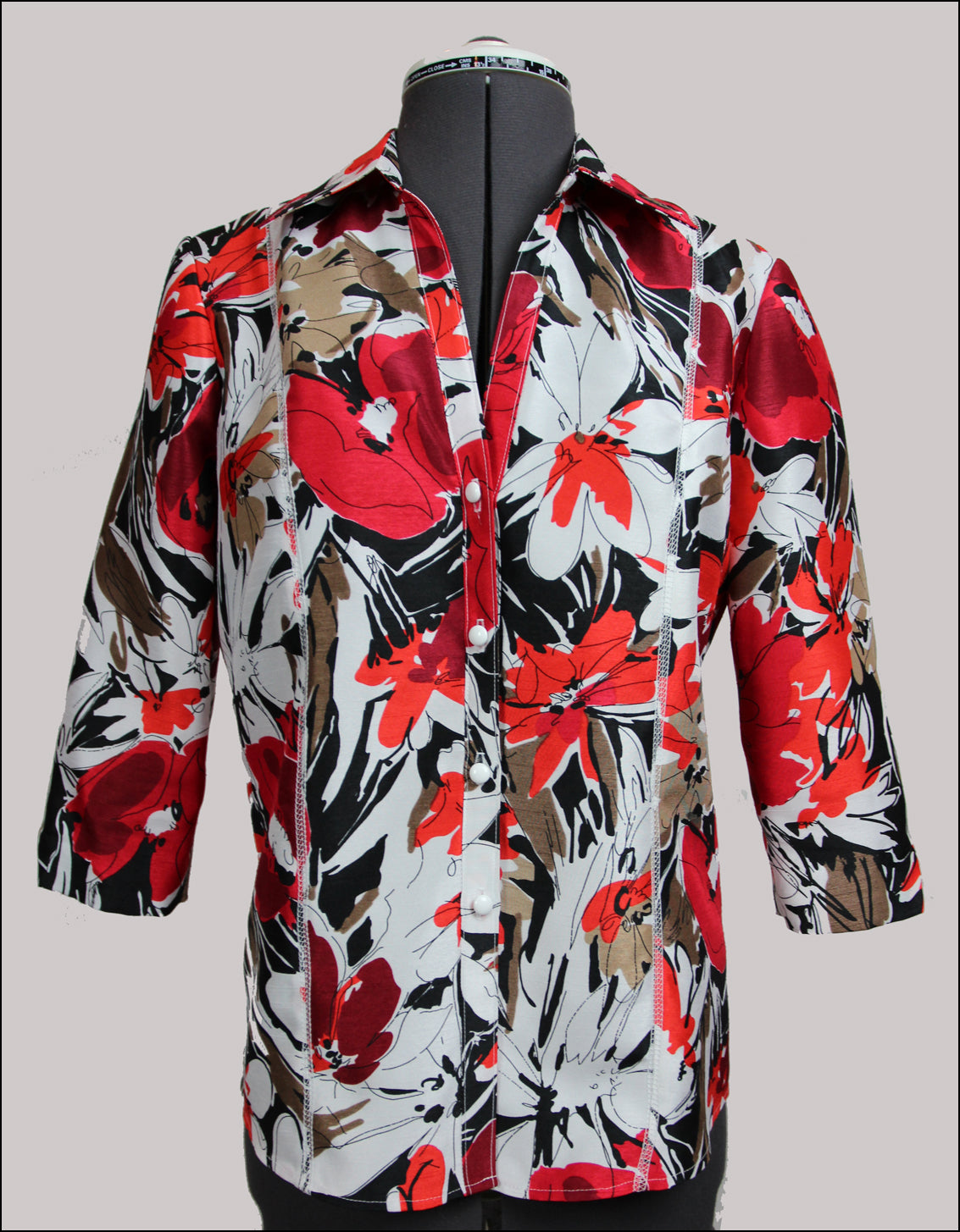 A digital sewing patter for a  3/4 sleeve shirt that buttons up the front with a collar.  The garment is on a mannequin. the fabric is large flowers of red orange and white 