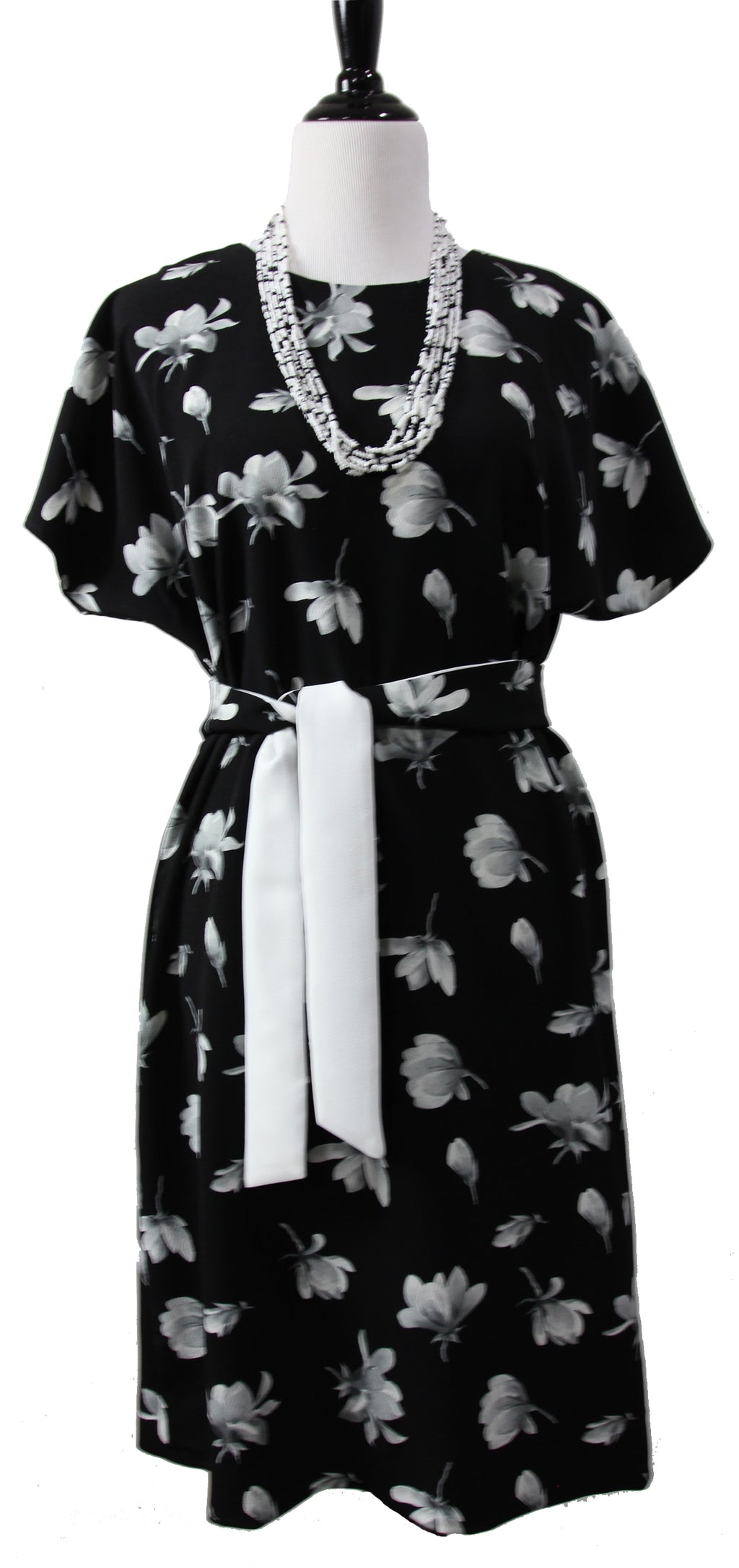 A close up of a short sleeve dress on a mannequin. it is made of black fabric with large white and gray flowers and a  gray beaded necklace and a long tie belt