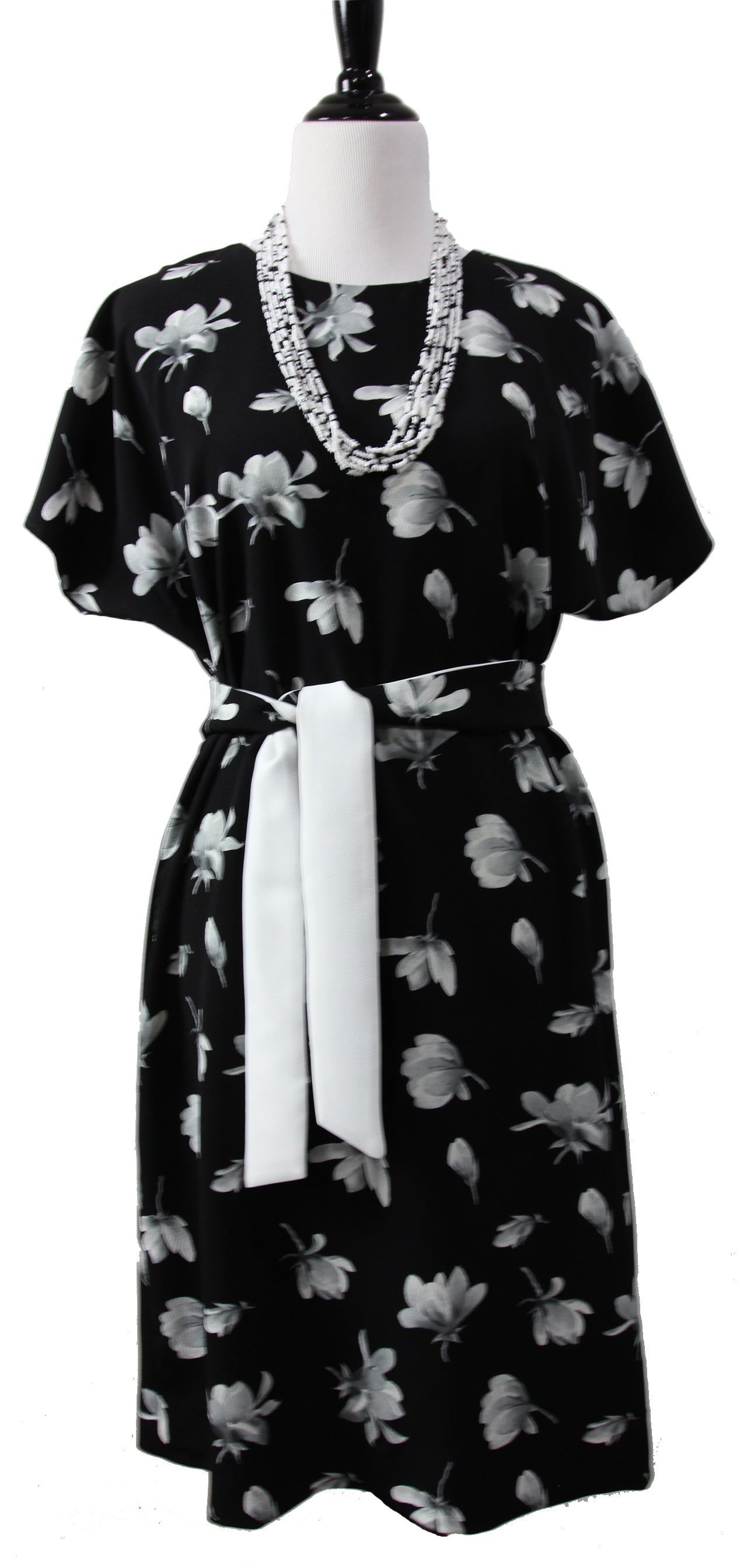 A close up of a short sleeve dress on a mannequin. it is made of black fabric with large white and gray flowers and a  gray beaded necklace and a long tie belt
