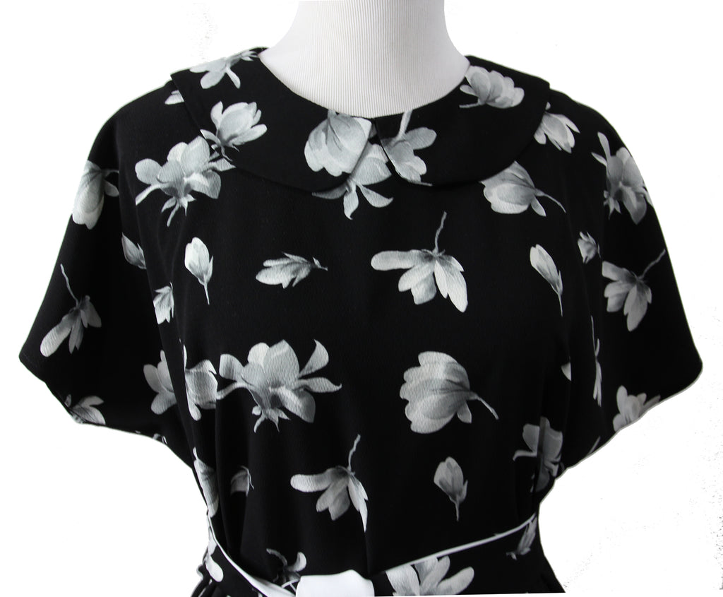 A close up of a short sleeve blouse on a mannequin. it is made of black fabric with large white and gray flowers and a rounded self fabric collar