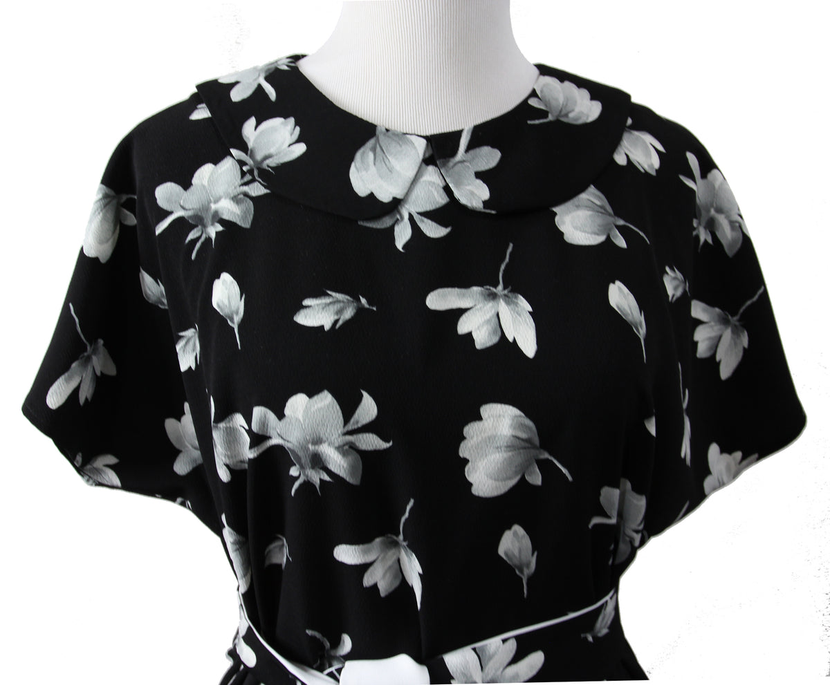 A close up of a short sleeve blouse on a mannequin. it is made of black fabric with large white and gray flowers and a rounded self fabric collar