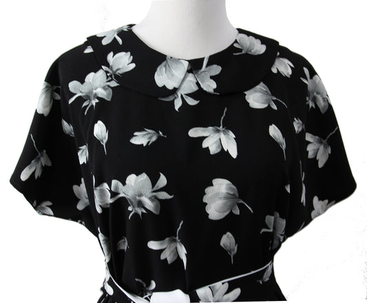 A close up of a short sleeve blouse on a mannequin. it is made of black fabric with large white and gray flowers and a rounded self fabric collar